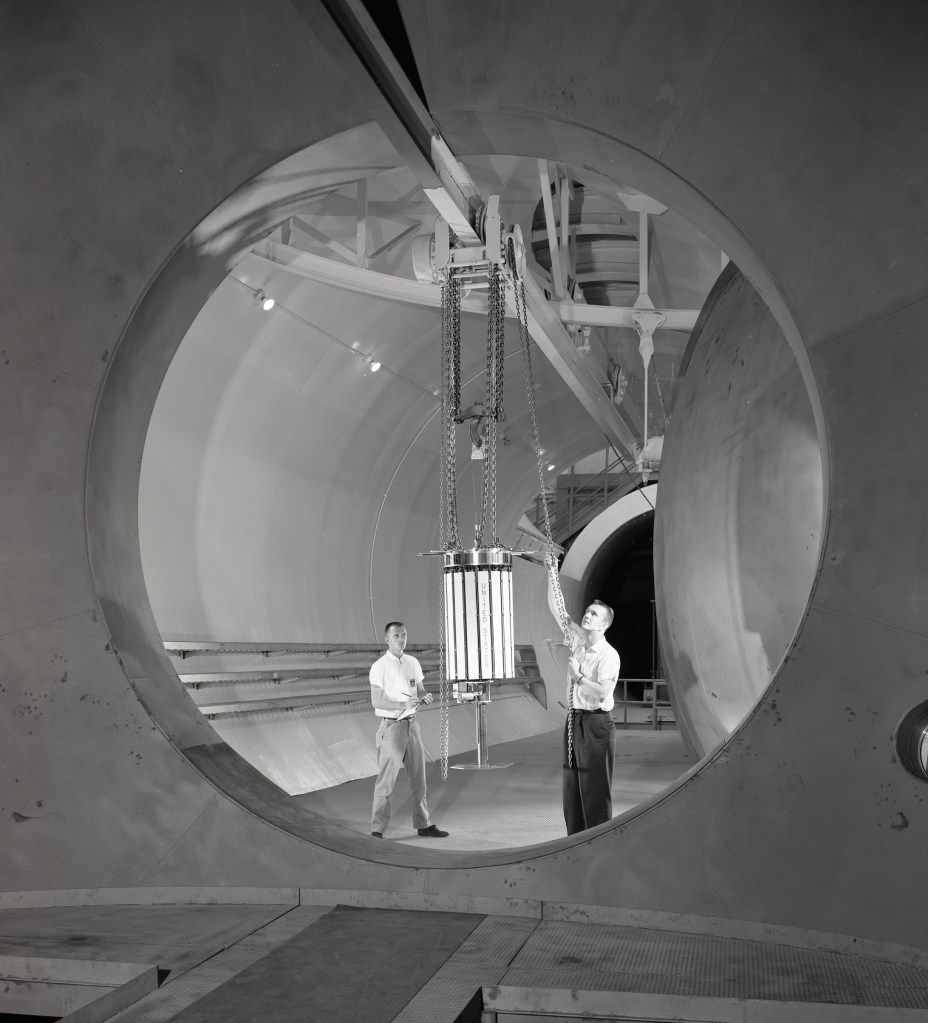 Two men move satellite with crane in chamber.