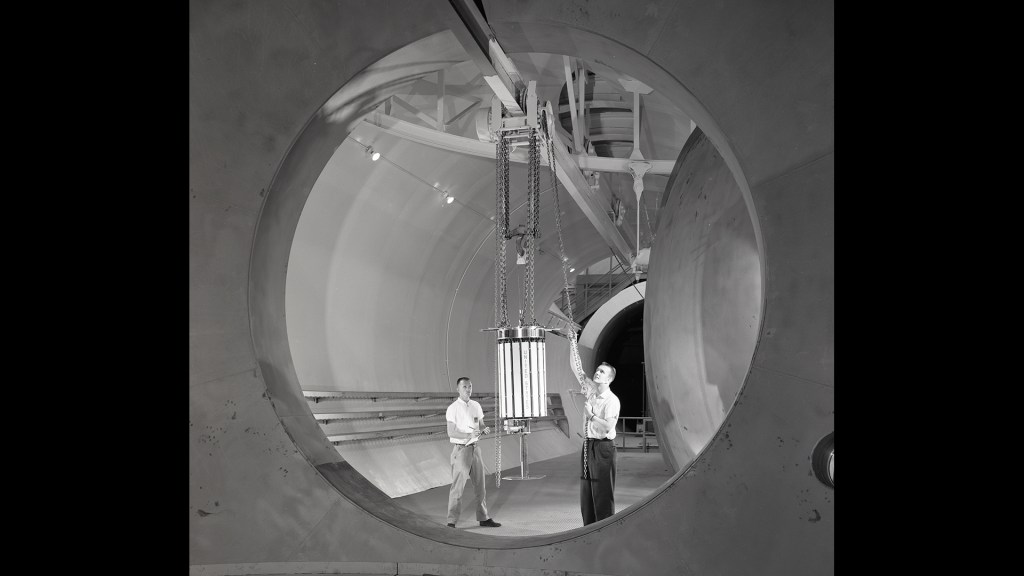 Two men move satellite with crane in chamber.
