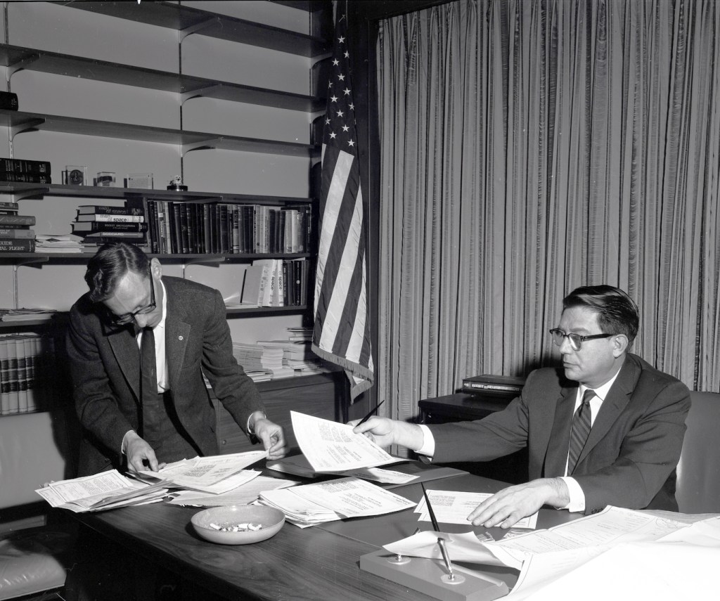 Man at desk handing paper to another person.