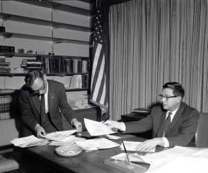 Man at desk handing paper to another person.