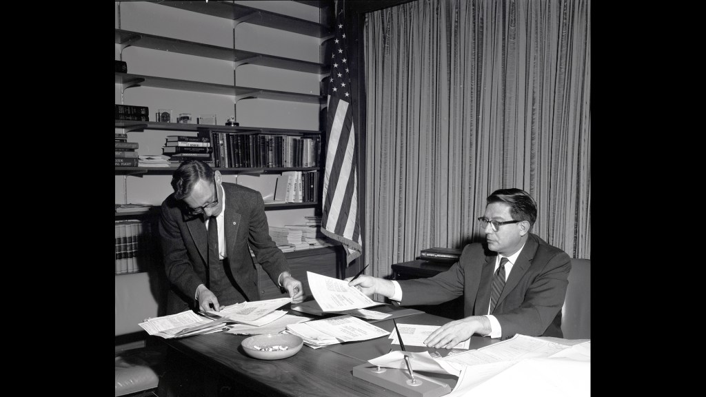 Man at desk handing paper to another person.