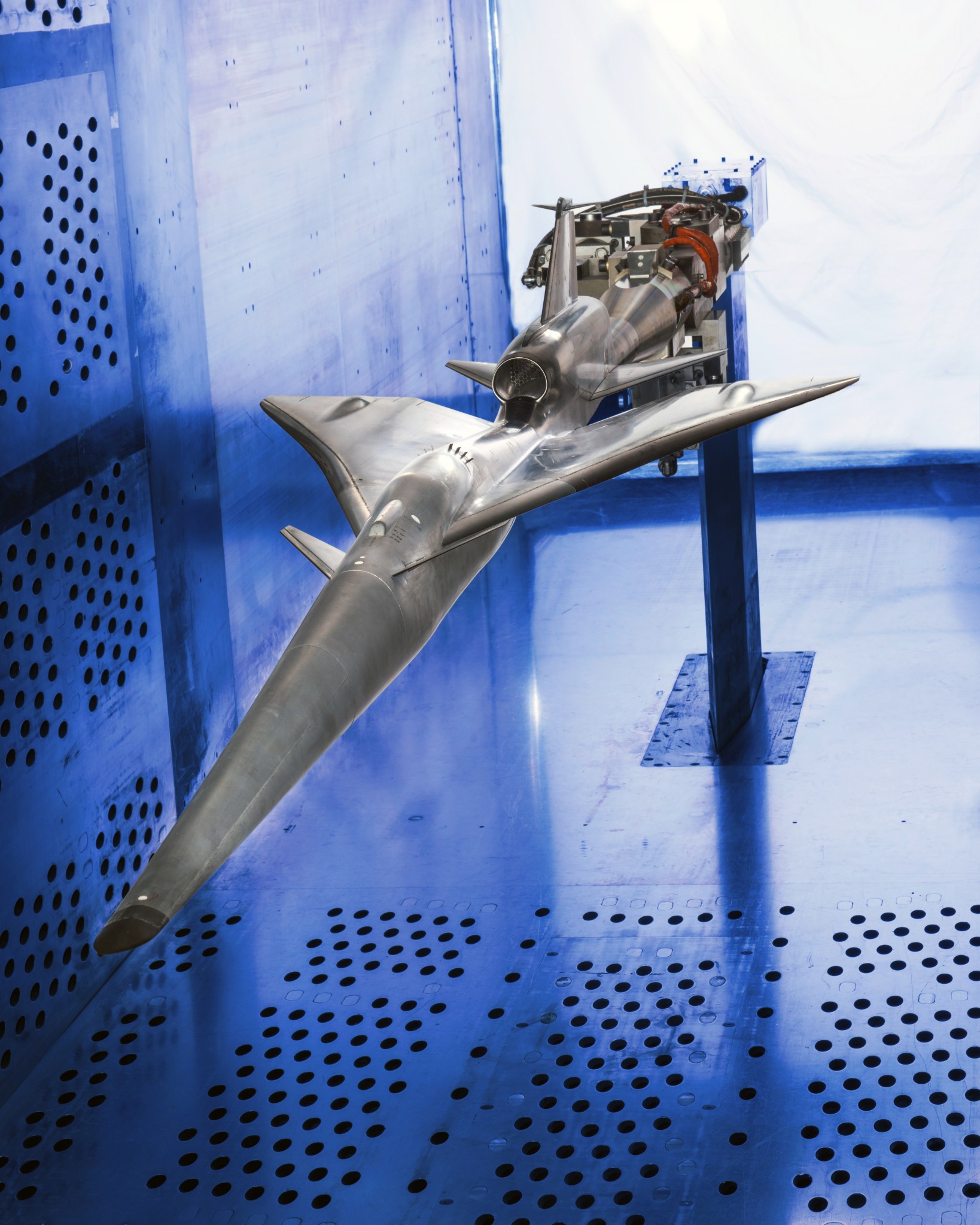 A metallic aircraft model mounted on a support inside a blue wind tunnel, surrounded by perforated walls and floor for aerodynamic testing.