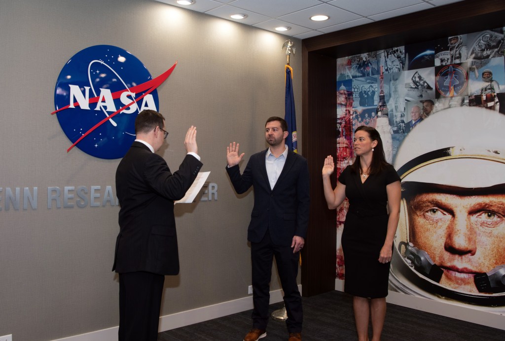 Man swearing in two people in front of John Glenn painting.