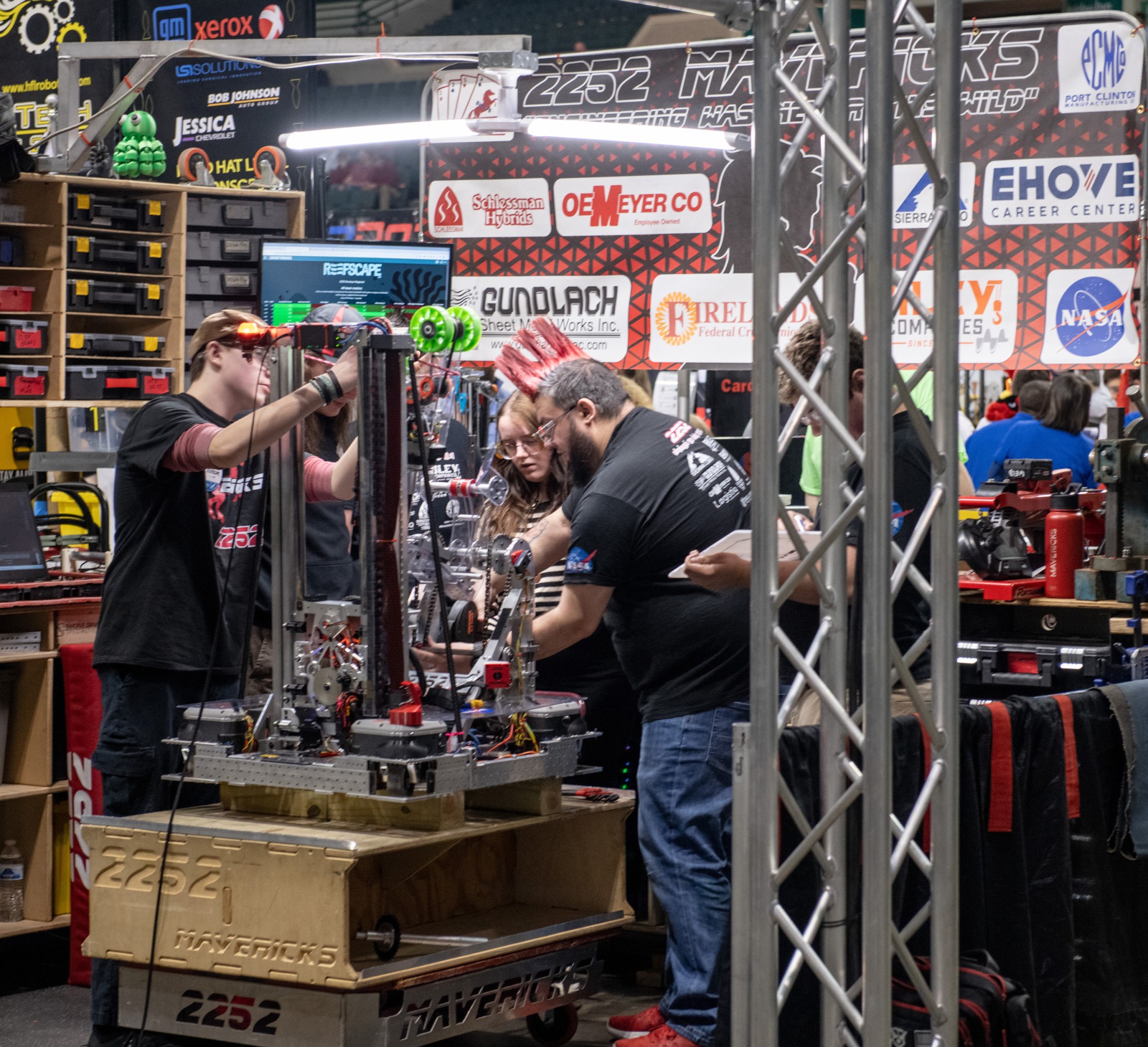 Four team members stand inside a small work area and gather around a robot as they adjust its parts prior to the game. Shelves of toolboxes and logos of company supporters surround them on two sides.