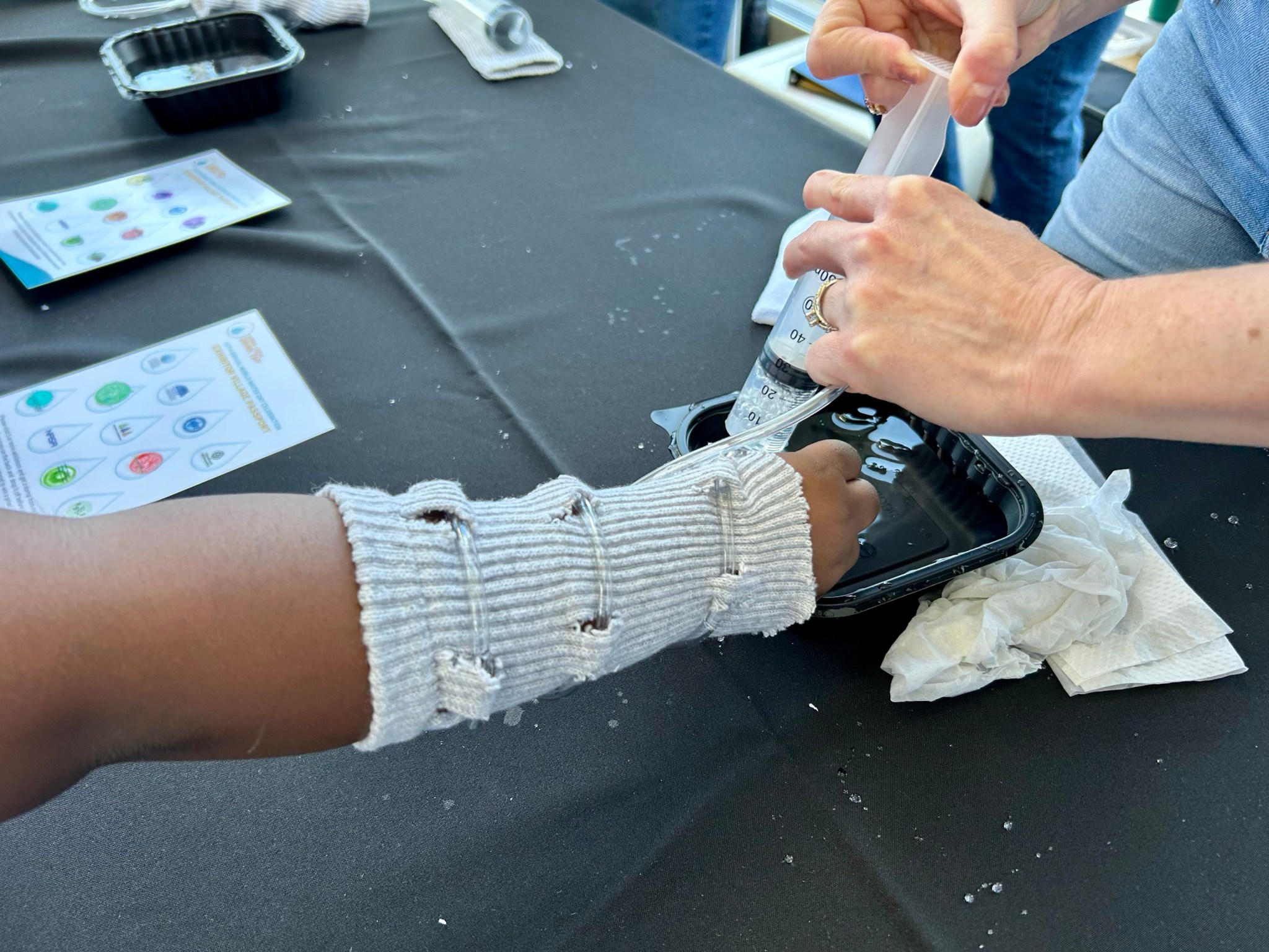 An up-close view of a student’s hand and forearm wearing the Liquid Cooling and Ventilation Garment. A NASA staff member uses a large syringe to dispense water into a tube that wraps around the garment.