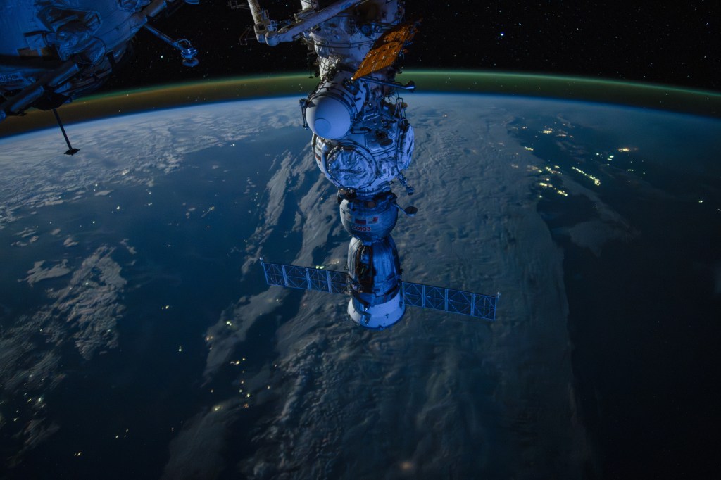 Looking aft from the cupola towards Soyuz over Southeast Asia at night. A sun about to rise from behind the camera (forward of space station) provides the light blue light to illuminate Soyuz.