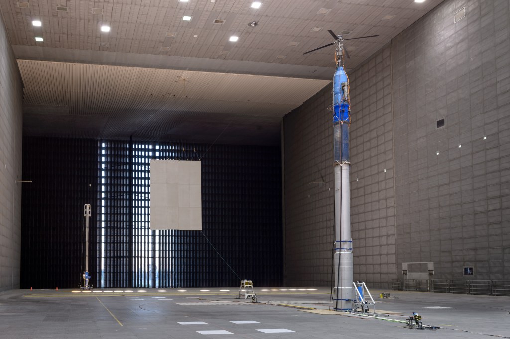 A metal tower with a four-bladed rotor on top and several small cameras looking up at it from the floor sits inside an expansive room that is part of a large wind tunnel.
