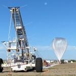 A scientific high-altitude balloon launch setup in a remote, open area. On the left is a specialized launch vehicle with large wheels and a tall, tower-like structure equipped with instruments and tethering equipment. On the right is a partially inflated white scientific balloon made of thin material, tethered and preparing for launch. The scene takes place under a clear blue sky with a visible moon, on a flat, open landscape. Orange safety cones mark the work area.