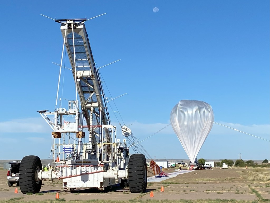 A scientific high-altitude balloon launch setup in a remote, open area. On the left is a specialized launch vehicle with large wheels and a tall, tower-like structure equipped with instruments and tethering equipment. On the right is a partially inflated white scientific balloon made of thin material, tethered and preparing for launch. The scene takes place under a clear blue sky with a visible moon, on a flat, open landscape. Orange safety cones mark the work area.