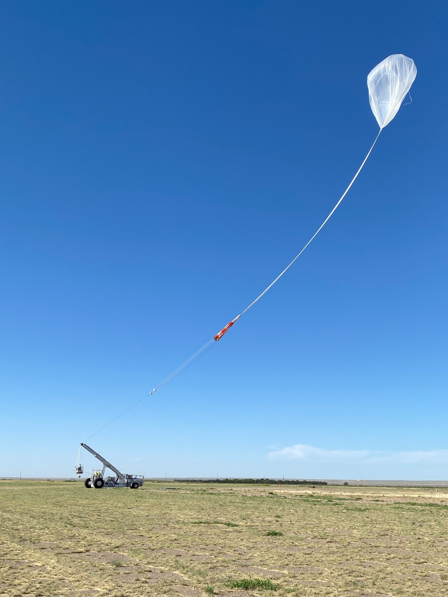 A scientific high-altitude balloon in flight, tethered to a launch vehicle on the ground. The translucent white balloon is high in a vivid blue sky, connected by a long tether line with a collapsed white and red striped parachute visible in the middle of the tether. At the bottom of the image, a small launch vehicle with a crane or winch system can be seen on a flat, grassy field.