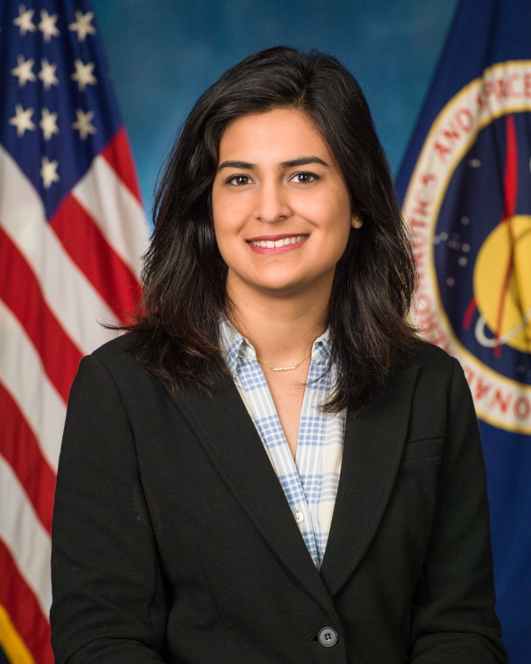 A woman in a dark suit with long dark hair poses with a smile in front of the American and the NASA flags.