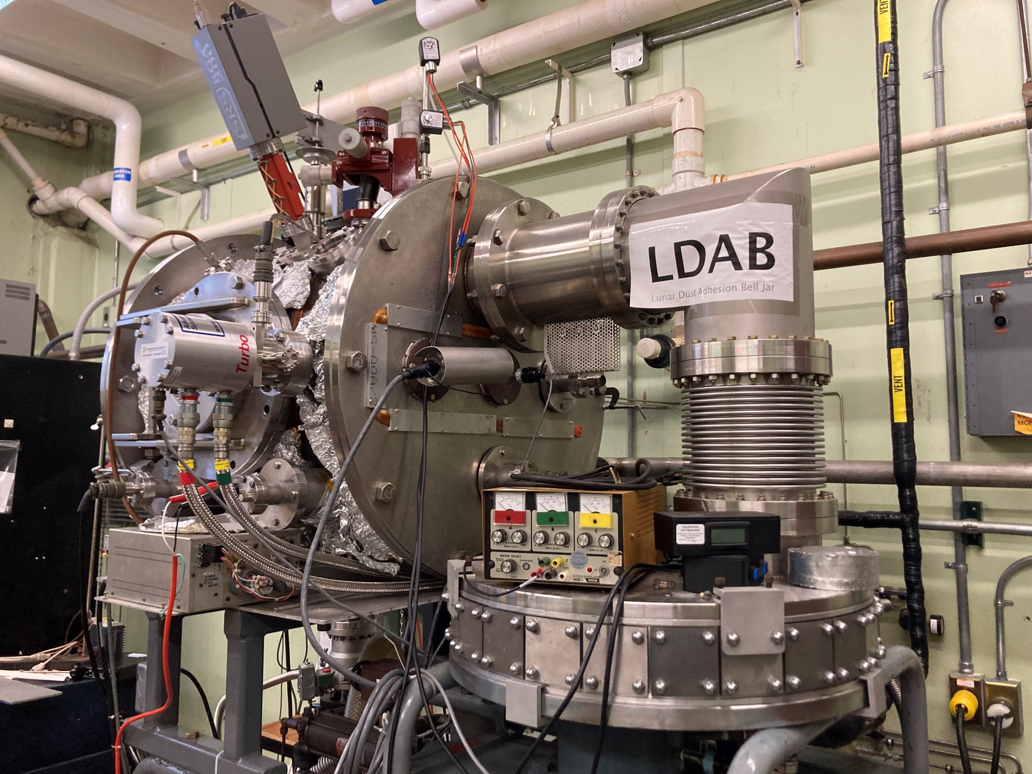 A vacuum chamber has a pipe exiting the chamber at a right angle, leading into a vacuum pump. Scientific instruments surround the chamber.