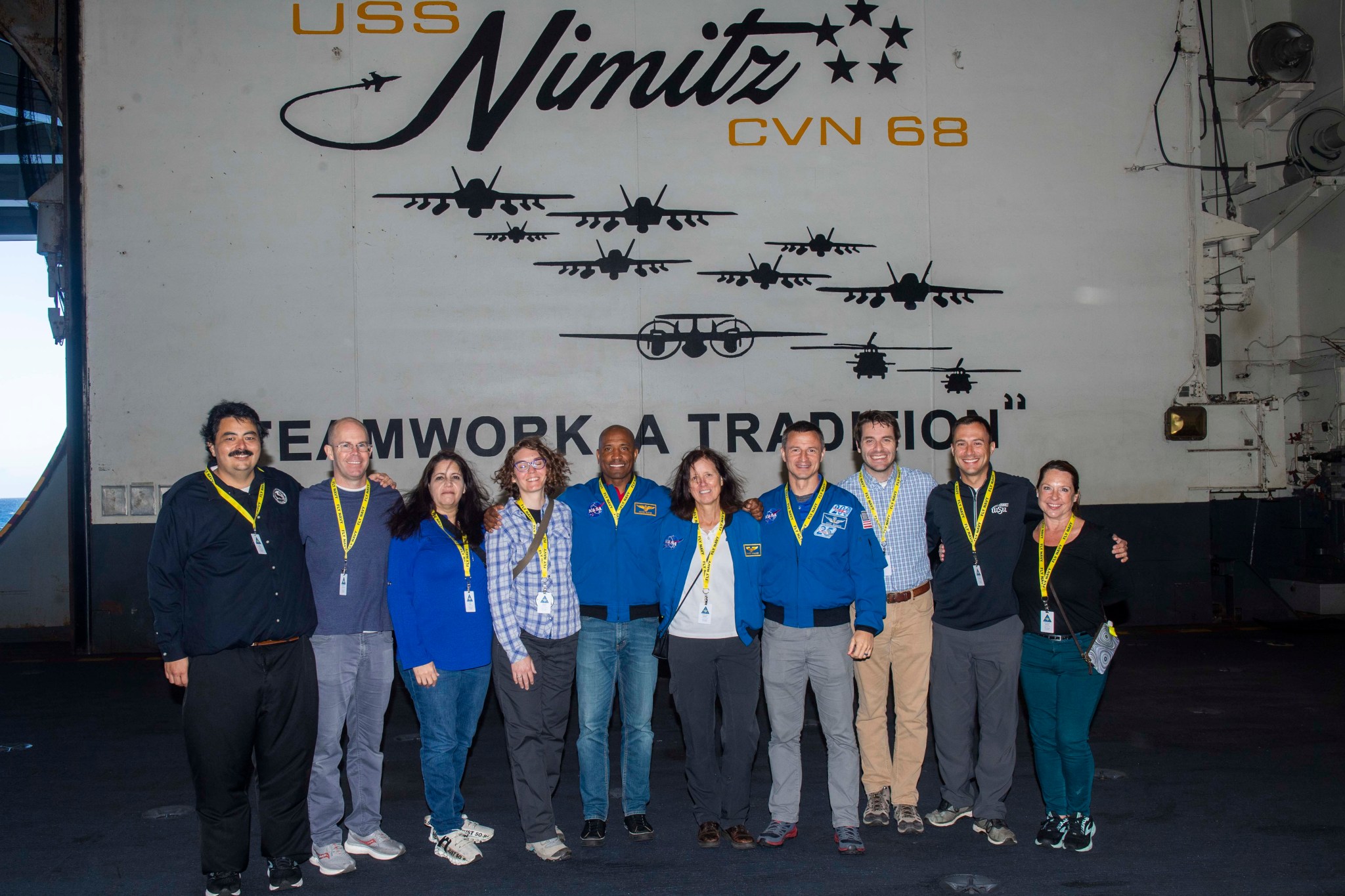 Ten people, four wearing blue flight suit jackets, pose in front of a building labeled, "USS Nimitz CVN 68."