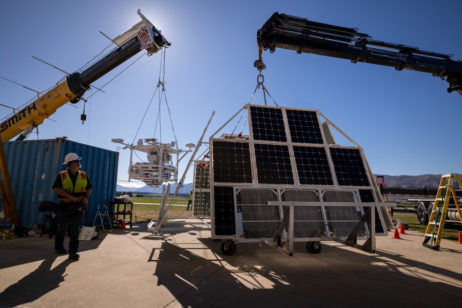 A large metal scientific instrument is suspended off the ground by a yellow crane. A black crane suspends black solar panels. A person stands in front of the instrument with a hard hat. It is daytime.