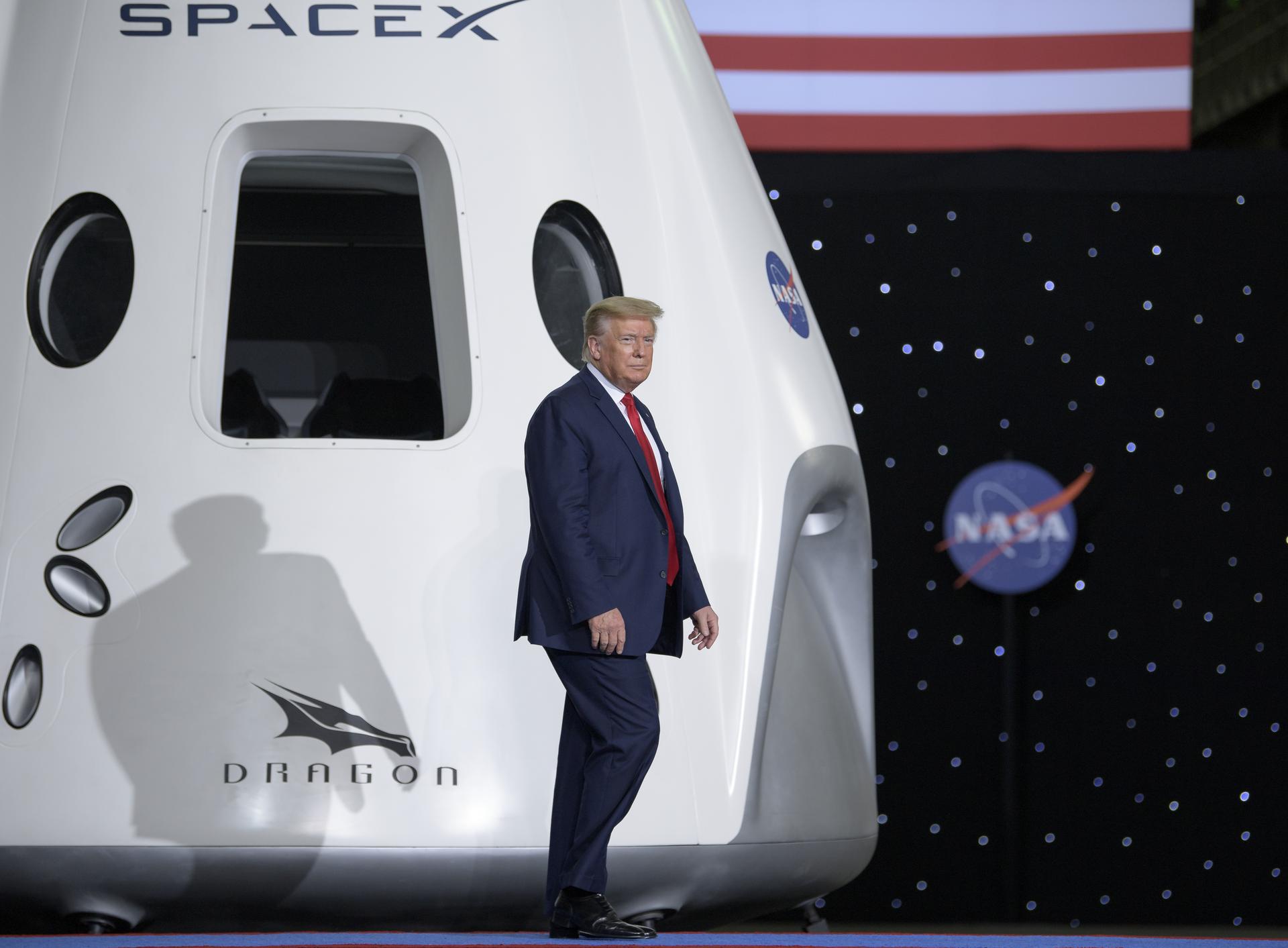 President Donald Trump walks onstage. Behind him is a white SpaceX Dragon crew spacecraft. It has a large rectangular window with two round windows on either side. The SpaceX, Dragon, and NASA logos can be seen on the spacecraft. In the background, the bottom of the United States flag (red and white stripes) are just visible at top, while the majority of the backdrop has small white lights, and another NASA "meatball" logo.