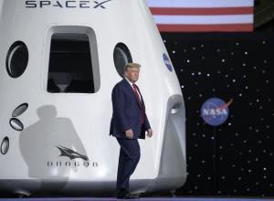 President Donald Trump walks onstage. Behind him is a white SpaceX Dragon crew spacecraft. It has a large rectangular window with two round windows on either side. The SpaceX, Dragon, and NASA logos can be seen on the spacecraft. In the background, the bottom of the United States flag (red and white stripes) are just visible at top, while the majority of the backdrop has small white lights, and another NASA "meatball" logo.