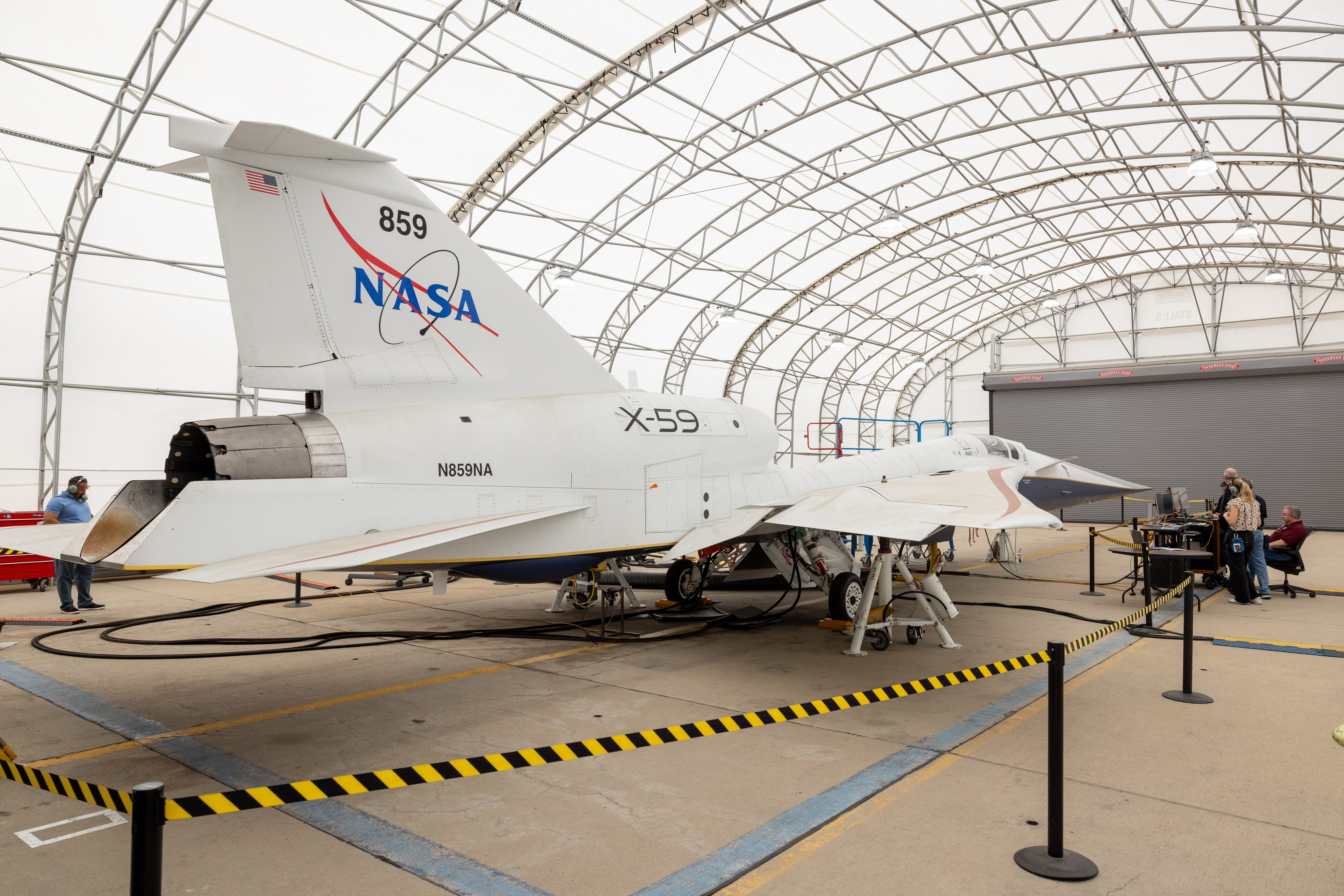 A white plane sits in a hangar that has a curved ceiling. The plane has markings on it including a NASA logo and "X-59." Several people are inside the hangar as well.