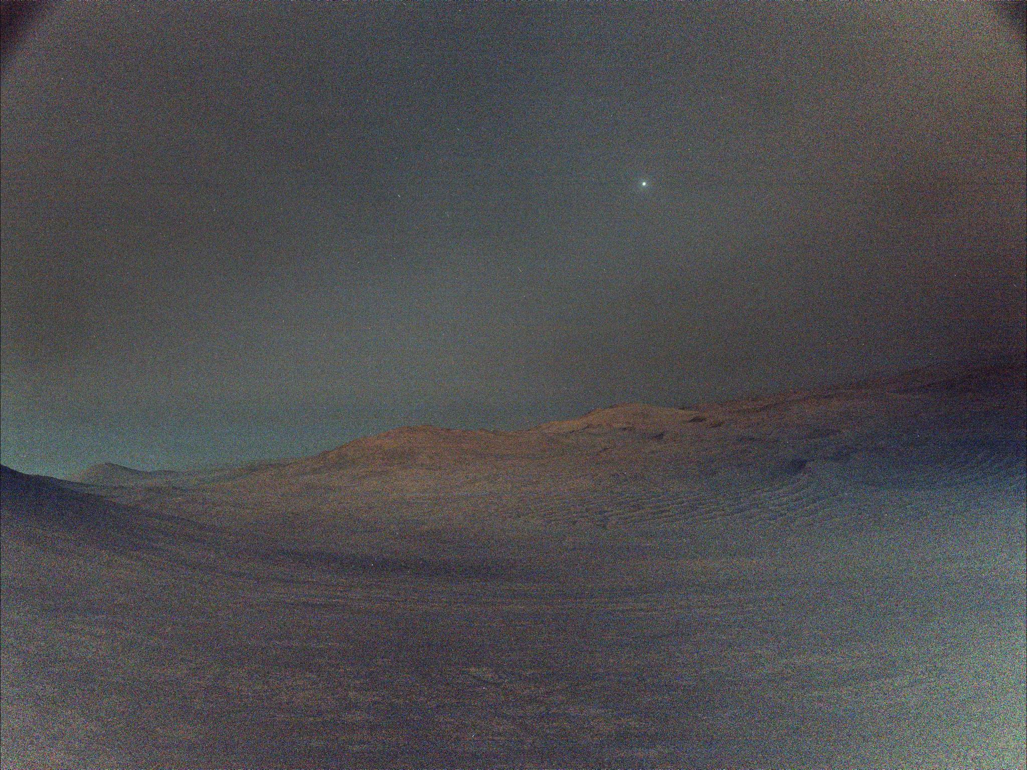 NASA's Perseverance rover captured this view of Deimos (the brightest spot in the sky), the smaller of Mars two moons, shining in the sky at 4:27 a.m. local time on March 1, 2025. The low light makes the view hazy, and the sky and ground both have a gray tint. In the distance, the crest of a crater shows Mars' signature rusty red color.