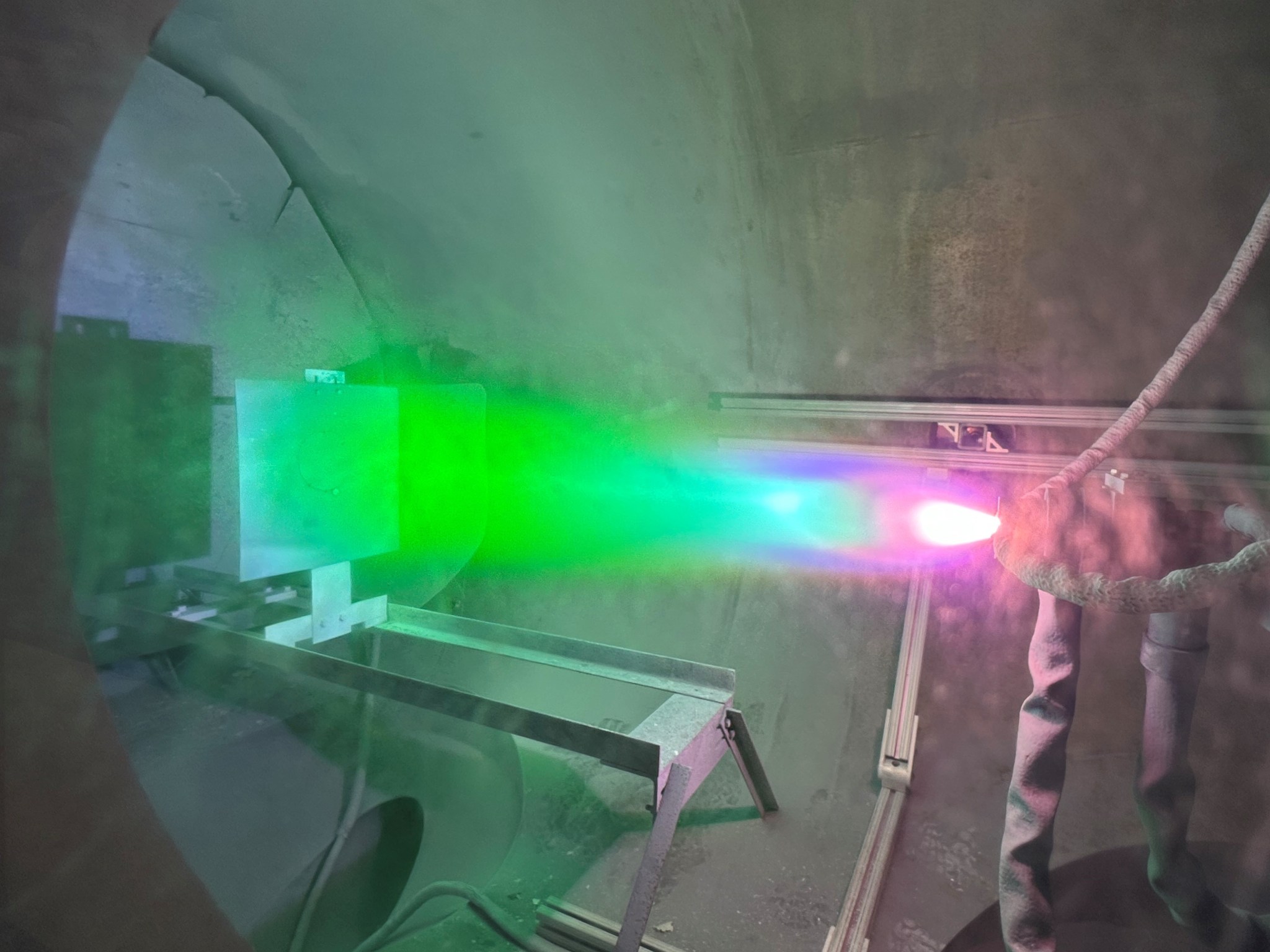 Inside a large chamber, a bright multicolored plasma sprays onto a surface.