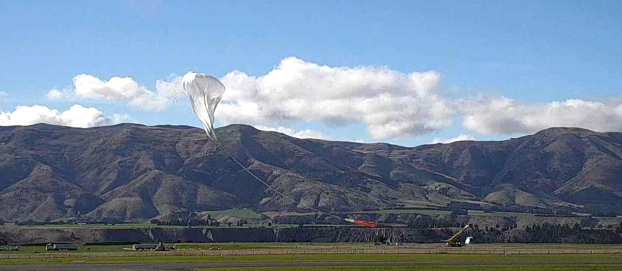 A large scientific balloon lifts from the ground. It’s attached by a long cable to a vehicle on the ground, which has a boxy scientific payload dangling from the right side on a long arm. The background has a mountain range with some clouds against a blue sky.