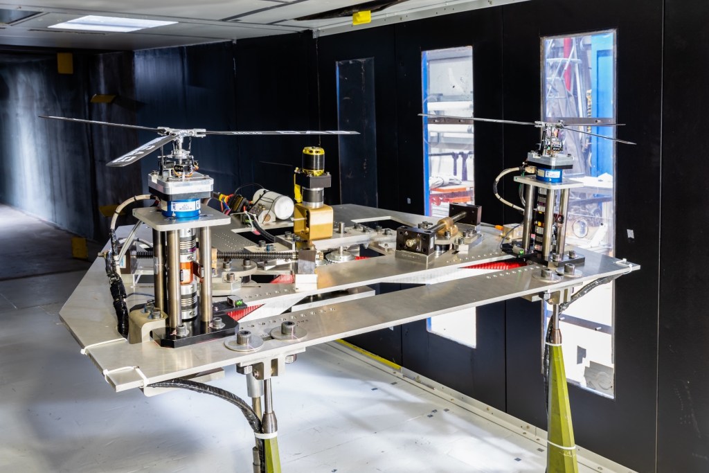 A helicopter-like model with two three-bladed rotors arranged side by side sits on a test stand in a wind tunnel.