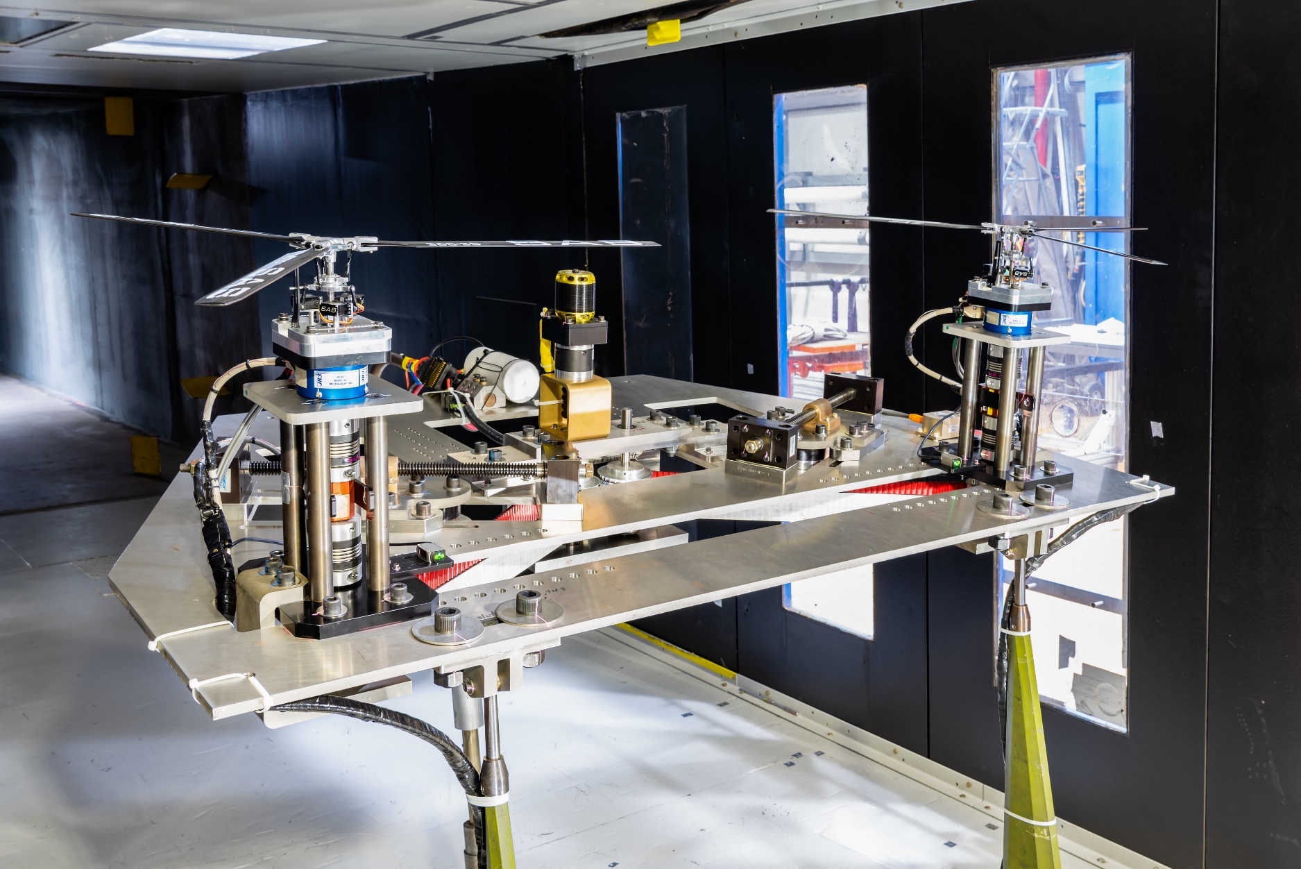 A helicopter-like model with two three-bladed rotors arranged side by side sits on a test stand in a wind tunnel.