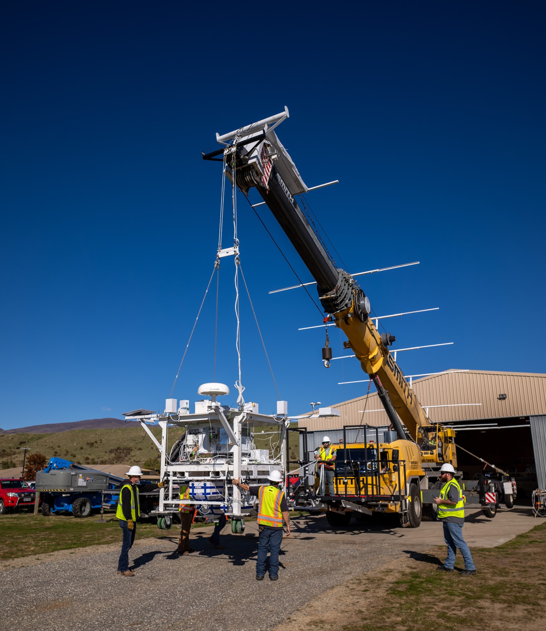 A large metal scientific instrument is suspended from a yellow crane with a hangar in the background.