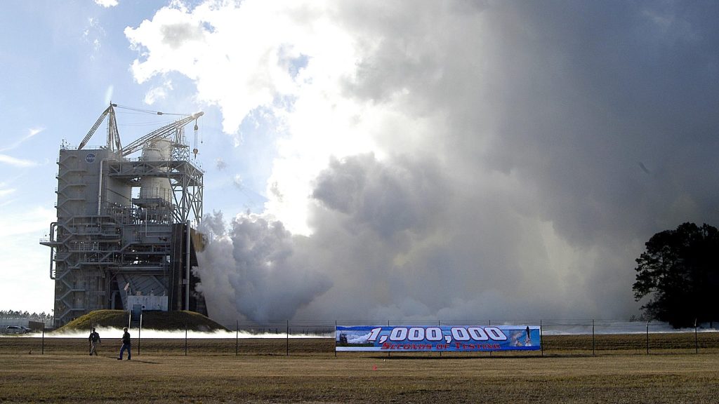 an 1 millionth second banner hangs on a fence to commemorate a successful test and flight operations of space shuttle main engines while vapor clouds erupt in the background