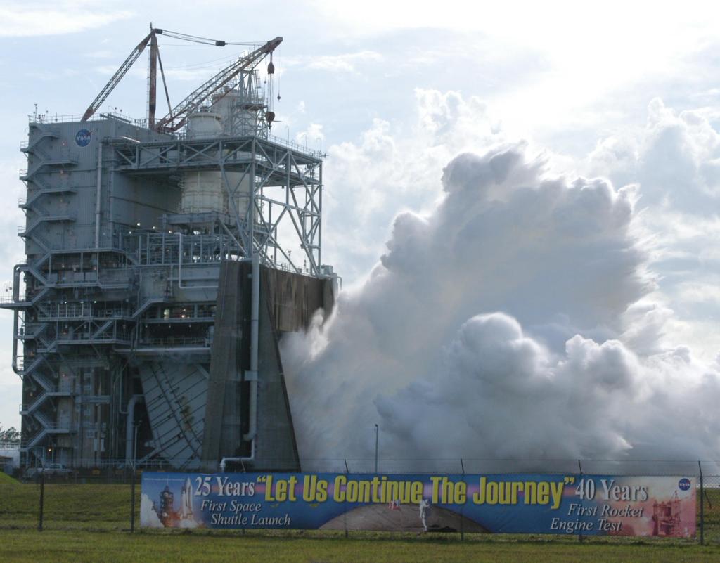 a banner hangs on a fence; in the background vapor erupts from A-2 Test Stand during the engine test