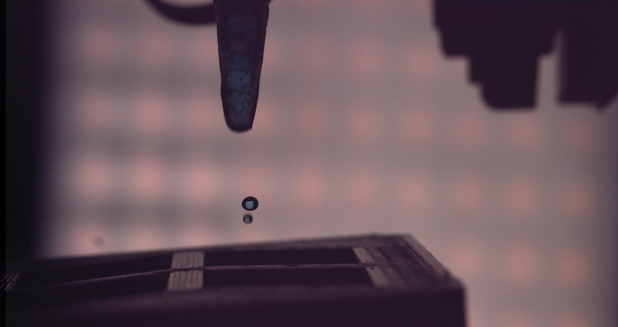 Closeup of the tip of a pipette with a droplet falling to a surface.