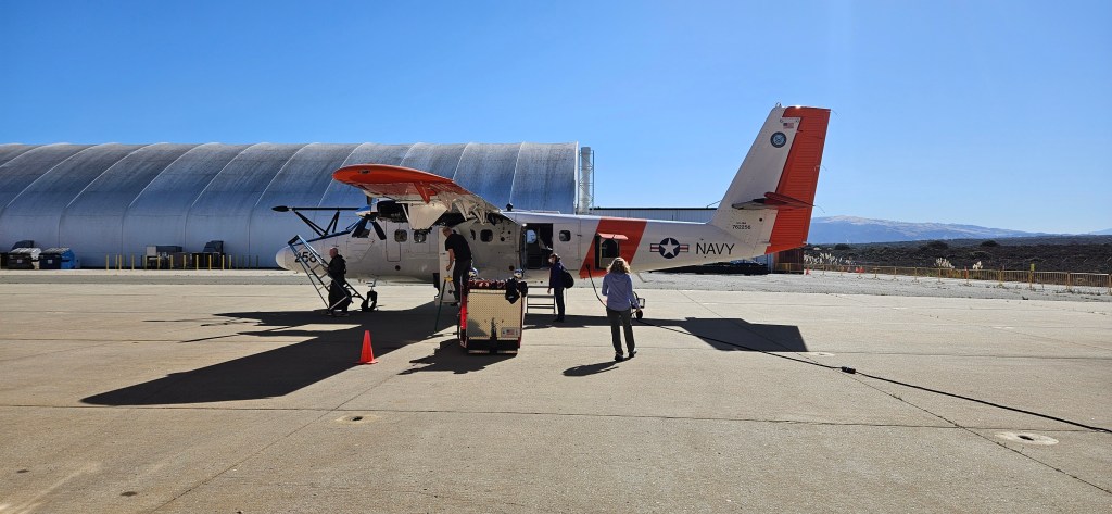 A small white plane with orange details sits on the tarmac with a silver hangar and blue sky behind it. A woman with blonde hair walks towards it.