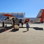 A small white plane with orange details sits on the tarmac with a silver hangar and blue sky behind it. A woman with blonde hair walks towards it.