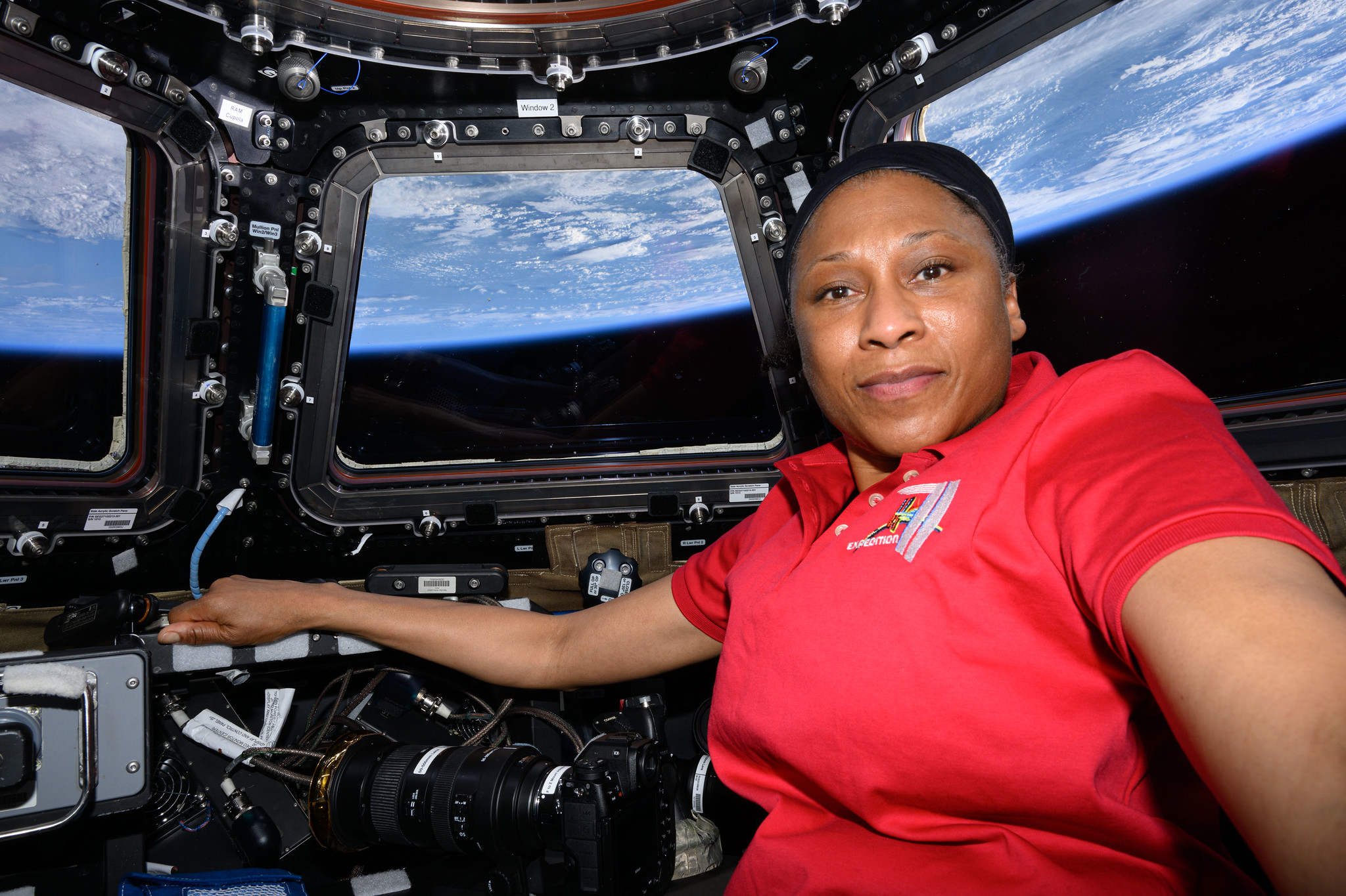 A black woman in a red shirt poses in the International Space Station cupola with the Earth pictured behind her.