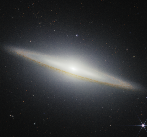 Image of a galaxy on the black background of space. The galaxy is a very oblong, brownish yellowish disk that extends from left to right at an angle (from about 10 o’clock to 5 o’clock). Mottled dark brown patches rim the edge of the disk and are particularly prominent where they cross directly in front of the galaxy. The galaxy’s center glows white and extends above and below the disk. There are different colored dots, distant galaxies, speckled among the black background of space surrounding the galaxy. At the bottom right, there is a particularly bright foreground star with Webb’s signature diffraction spikes.