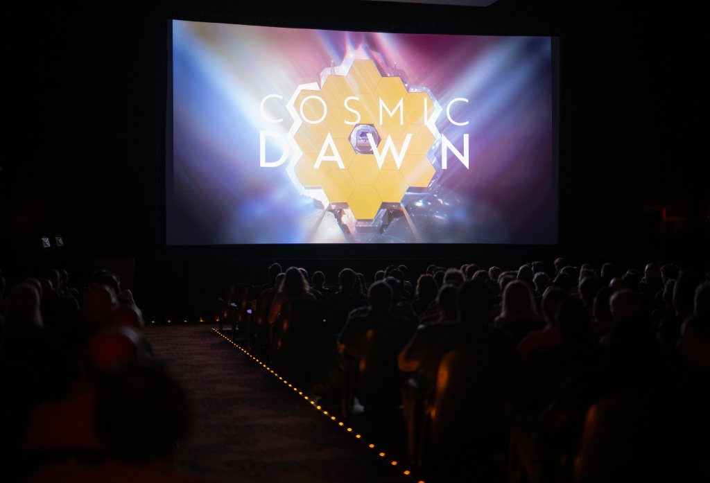 People sit in a dark theater with a bright movie screen in the front. The words "Cosmic Dawn" with the James Webb Space Telescope primary mirrors behind it.