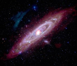 A rosy pink spiral galaxy is positioned diagonally in this picture. The flat disk shape has spiraling arms, and at center, a bright core. All over the image are other galaxies that look like specks of multicolored glitter.