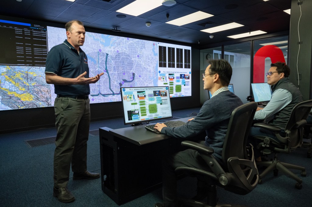 A NASA researcher presents information on a large screen at the front of the room to an audience. Two audience members can be seen, each behind a computer station.
