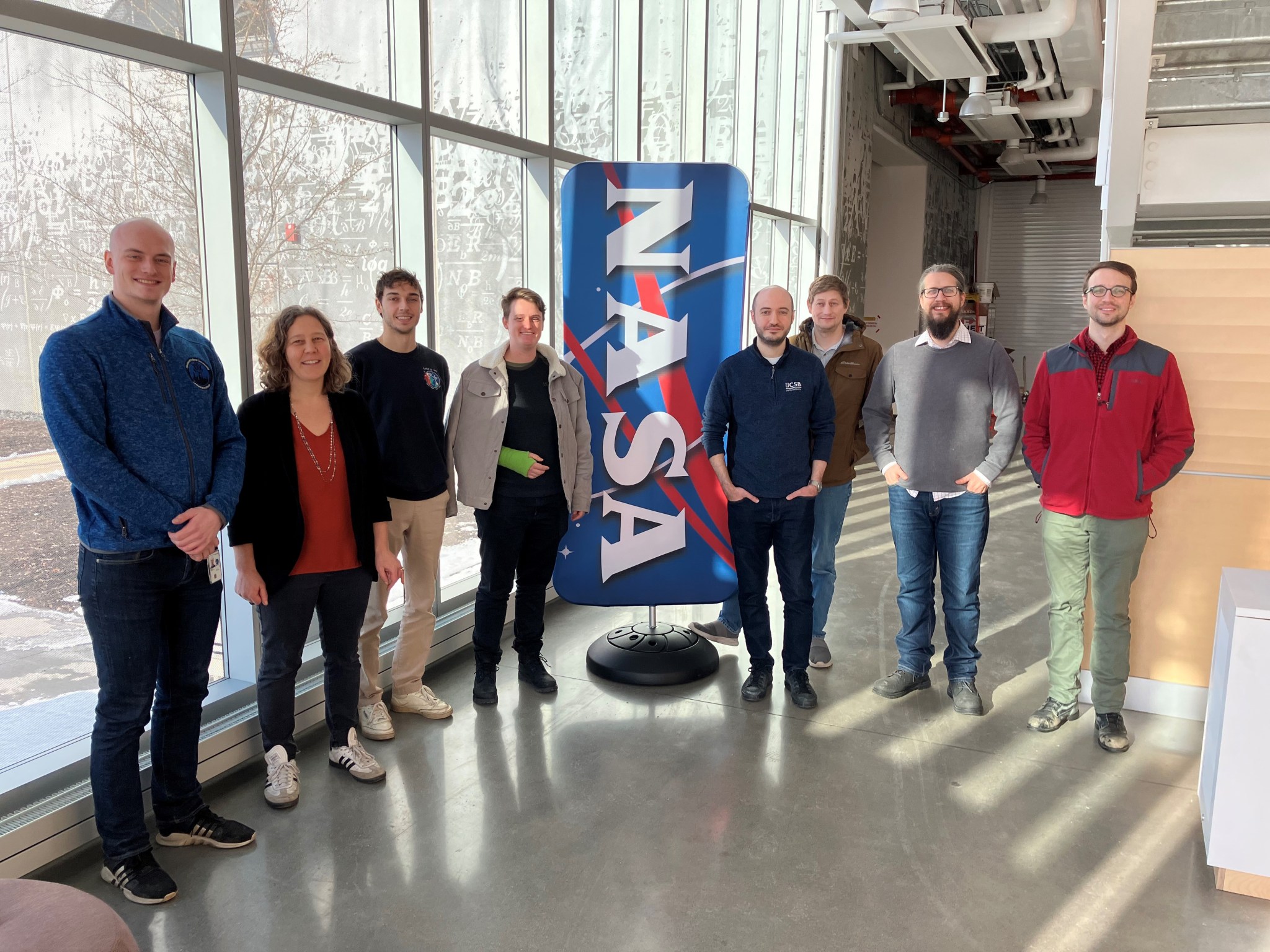 A group of eight individuals from the RealTOR research team stand in a line posing for a group photo. The team is smiling standing in front of a bright blue NASA banner, in a large room with tall windows that let in natural lighting.