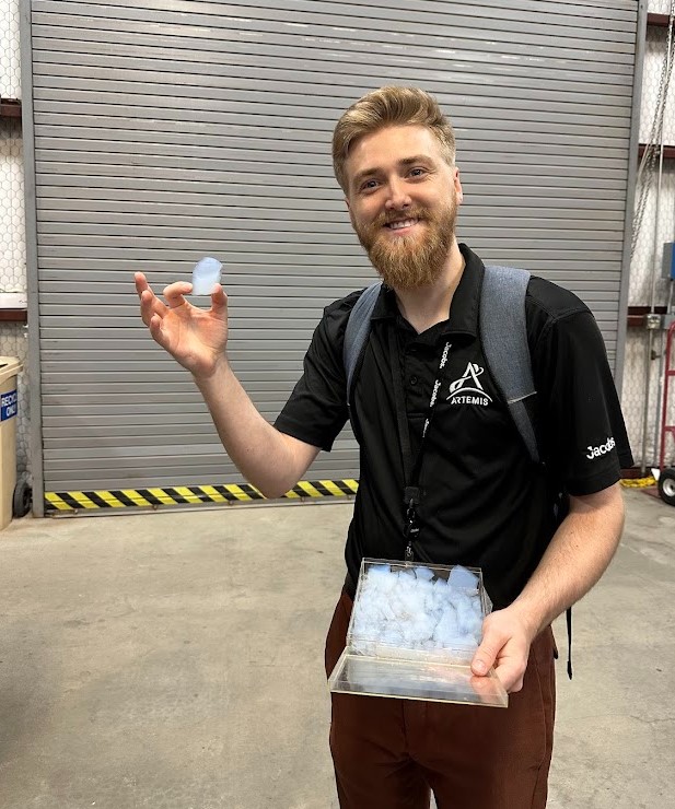 A man wearing an Artemis polo shirt and brown pants holds a piece of Aerogel in his right hand and a container of other Aerogel pieces in his left hand.