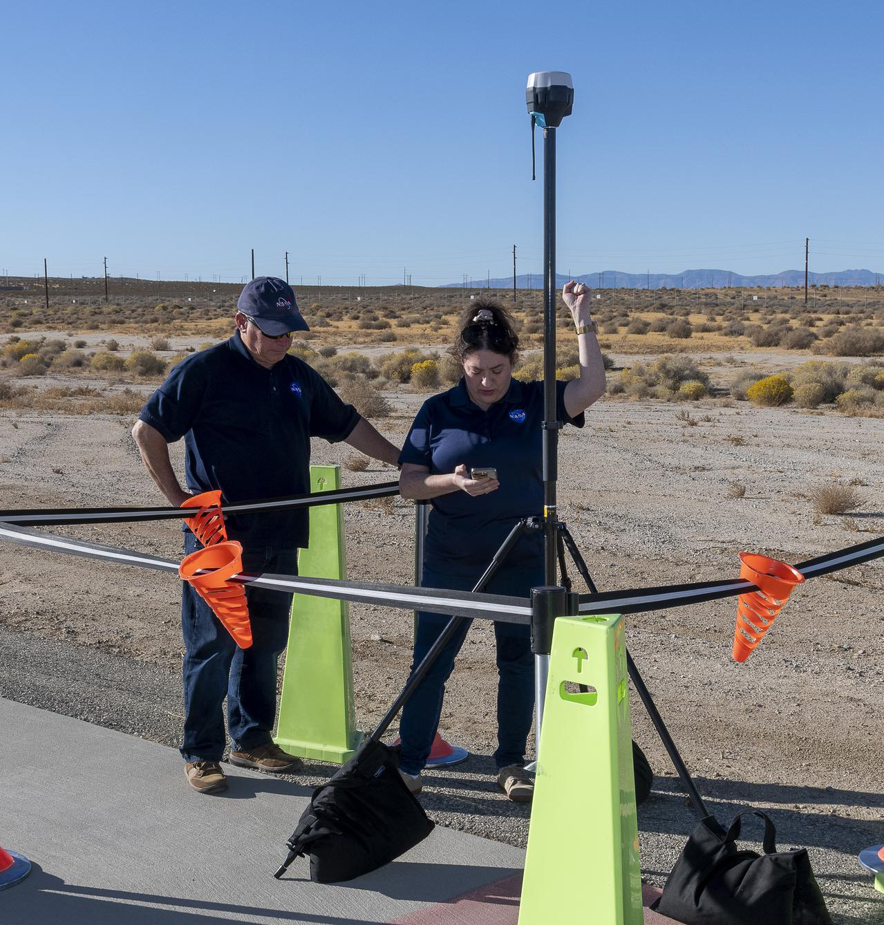 NASA Pathways intern, Saré Culbertson, right, wearing a dark blue shirt with a NASA meatball logo, verifies the coordinates for an Emlid Reach RS2+ receiver mounted on a black metal tripod. The equipment is cordoned off with metal stands and black tape. Bright neon green cones mark the corners and orange plastic triangular cups hang from the center of the black tape. NASA operations engineer stands to the right of Culbertson, observing the validation data on the test equipment in her right hand. They are surrounded by desert scrub and sand at NASA’s Armstrong Flight Research Center in Edwards, California on November 7, 2024.