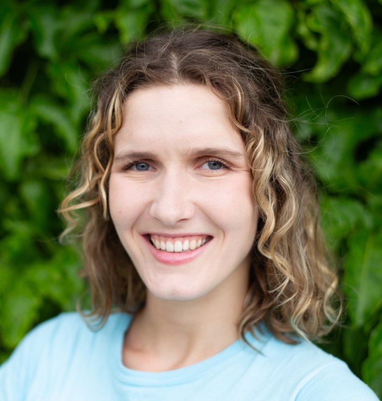 Portrait image of Angelique Ray wearing a light blue shirt standing in front of a green bush background.
