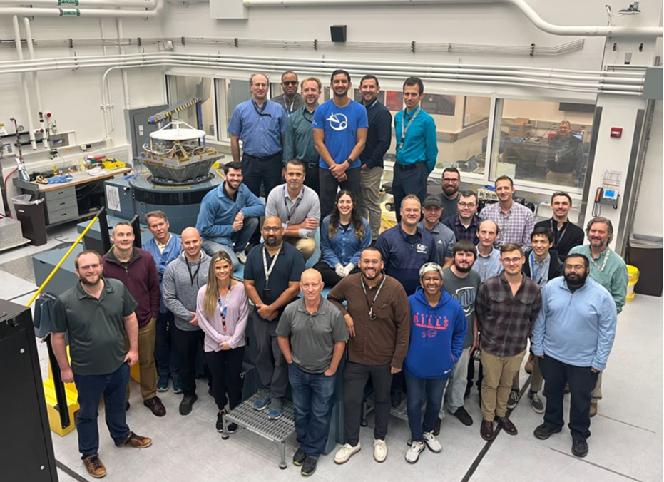 A group of thirty smiling engineers and technicians in casual and work attire stand inside the Johns Hopkins University Applied Physics Laboratory. To the left, the Polylingual Experimental Terminal (PExT) payload — featuring a white antenna dish and angular wired metal hardware — is mounted on a circular vibration table. The lab is a white, windowed cleanroom with gray flooring, white piping, test benches, and space integration equipment.