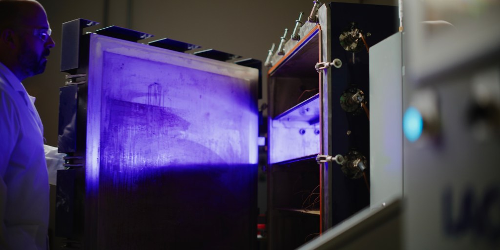 Person wearing lab coat holds open door to metal chamber with purple glow from inside.