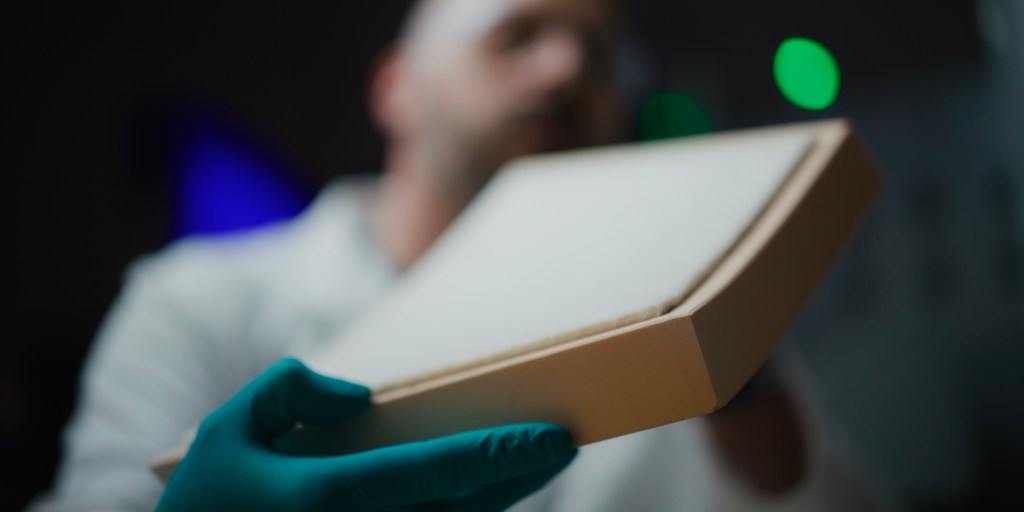 Person wearing lab coat, protective glasses and gloves holds curved thermal protection system tile.