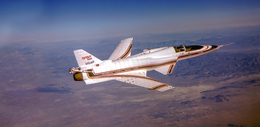 A one-person white jet with forward-swept wings flies high above a mountainous desert.