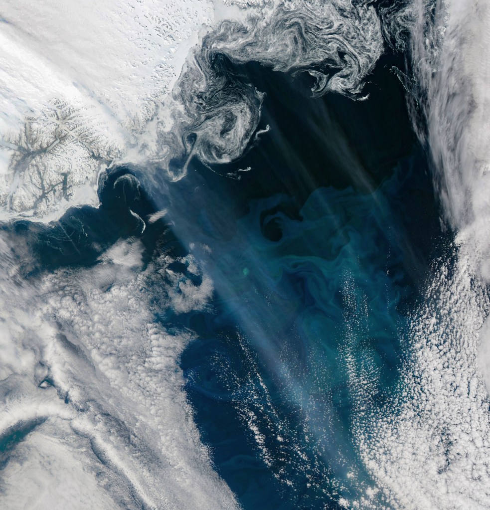 A satellite image shows a dark blue ocean with swirls of a slightly lighter blue. Textured white clouds encroach from each corner, making the the view of the ocean a rough diamond shape.
