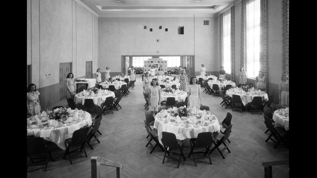Women standing by tables in auditorium.