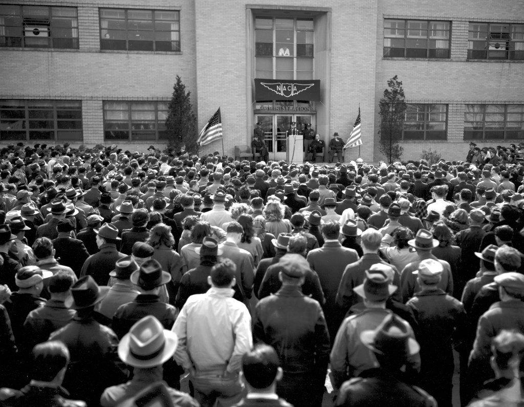 Group of people outside building listing to speaker.