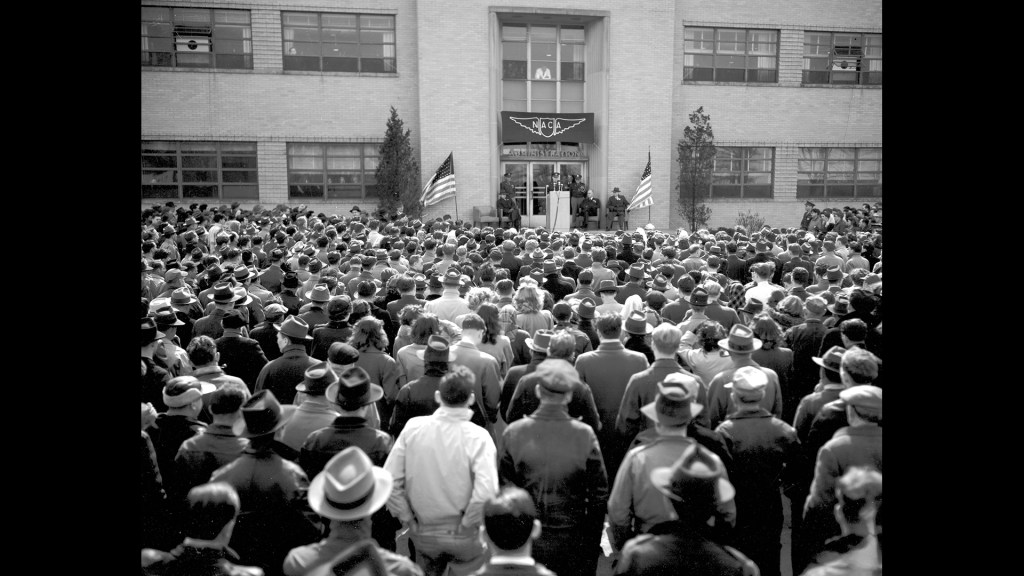 Group of people outside building listing to speaker.