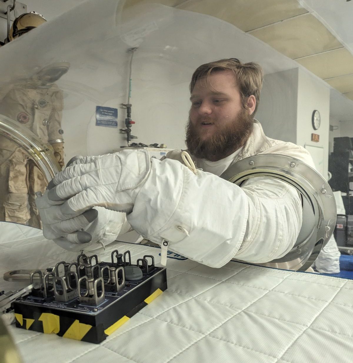 A man wearing a space suit glove works with tools inside of a glovebox.